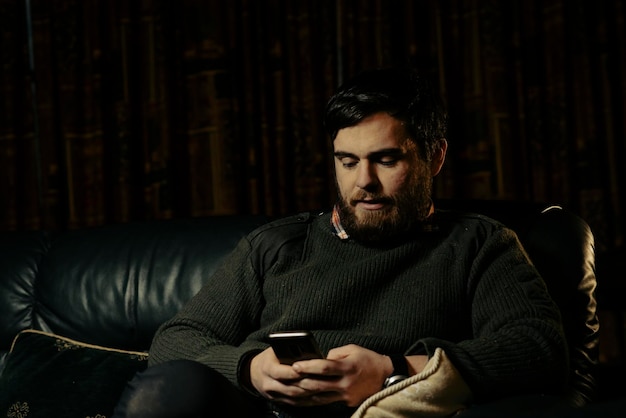 Handsome man looking at mobile phone while sitting on sofa in\
sweater in winter