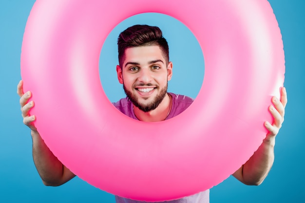 Handsome man looking through pink rubber ring for swimming isolated over blue