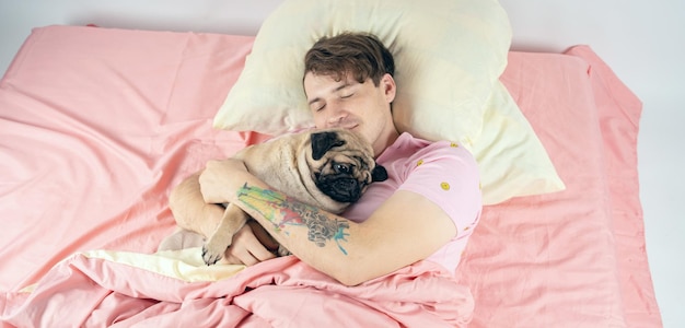 Handsome man lying with pug at bed Young man in a bed under a rug with his dog in an embrace Man with dog lying down in the bed in a morning and looking in to a camera