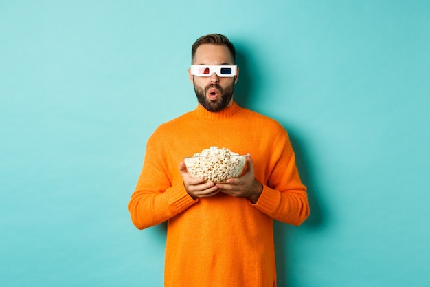 Handsome man in orange sweater and 3d glasses watching movies thrilled, holding popcorn, looking amazed, standing over blue background.