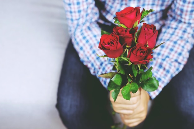 ソファに座っているハンサムな男性の格子縞のシャツが赤いバラの花と美しい花束を差し出す