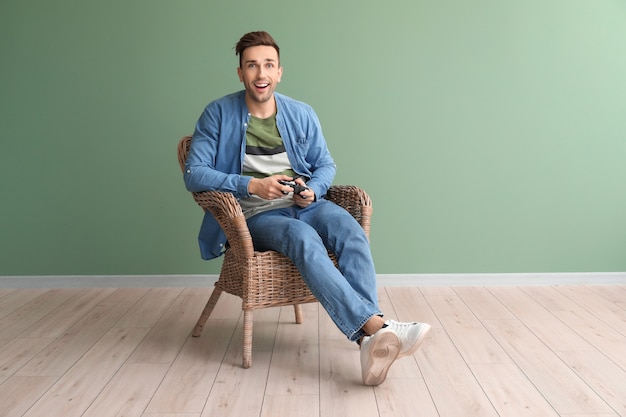 Handsome man playing video games while sitting in armchair near color wall