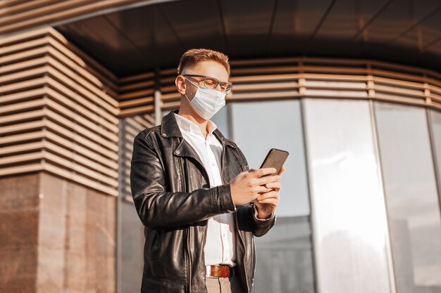 Handsome man in a protective mask on his face with glasses with a smartphone on the street of a big city. Businessman talking on the phone on urban background