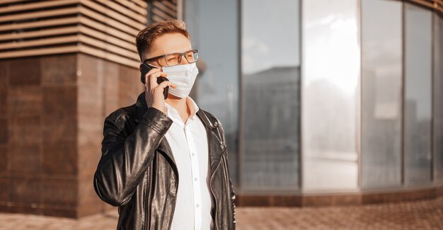 Handsome man in a protective mask on his face with glasses with a smartphone on the street of a big city. Businessman talking on the phone on urban background