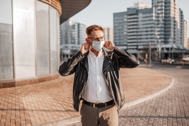 Красивый мужчина, надевающий медицинскую защитную маску на лицо в очках на улице большого города на городском фоне