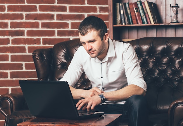 A handsome man reads information in the laptop on the background of a shelf with books.