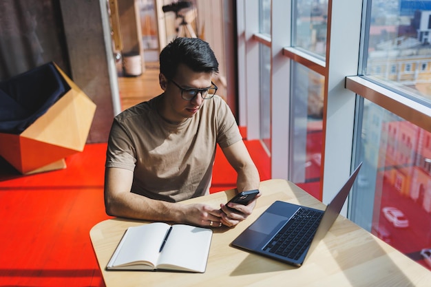 Handsome man in red casual tshirt writing a message on mobile phone while sitting near glass window at table with laptop working remotely in cafe