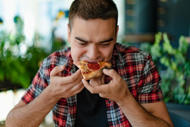 Handsome man in a red plaid shirt is eats a pizza at a cafe Hungry man eats a slice of hot pizza with cheese sausage and sauce Closeup