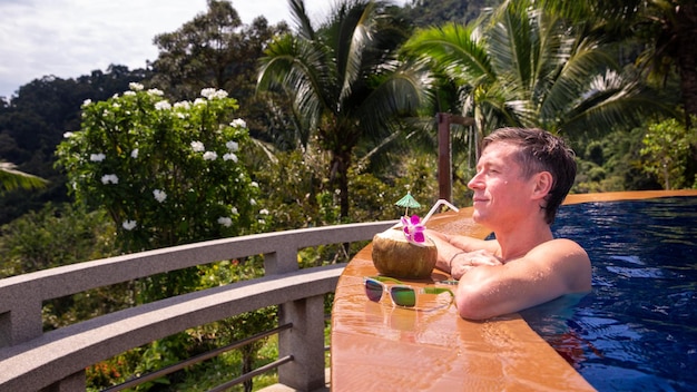 Handsome man relaxes by the outdoor pool and drinks a coconut with palm trees in the background Summer concept