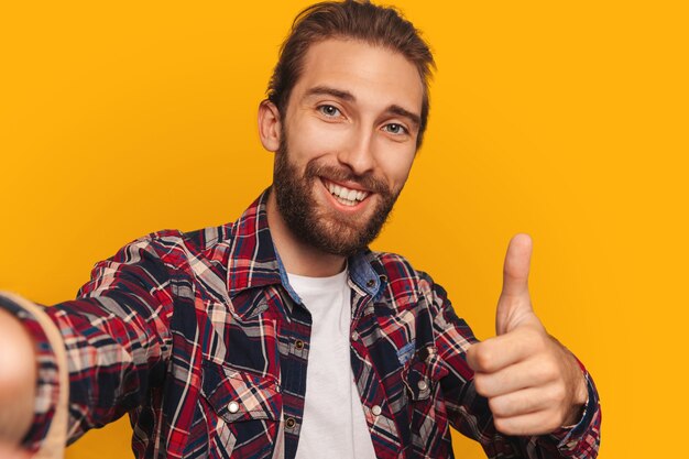 Handsome man in a shirt with a beard takes pictures of himself, smiles and shows a thumb up