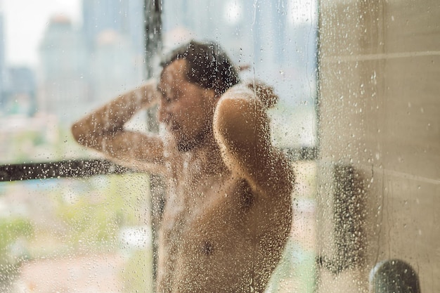 Handsome man in the shower behind glass with drops on the background of a window with a panoramic view of the city