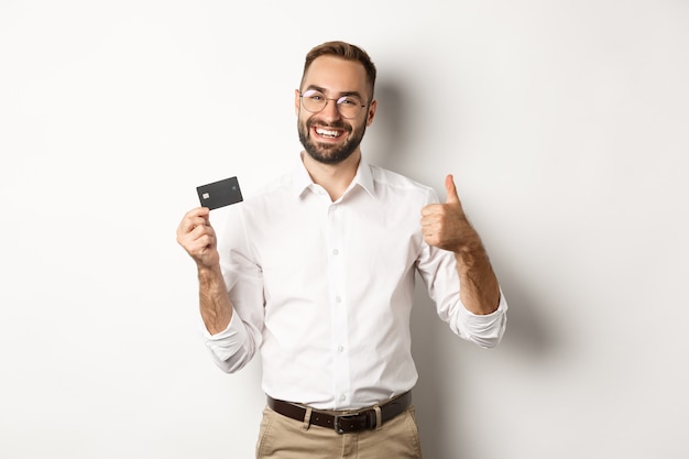 Красивый мужчина показывает свою кредитную карту и большой палец вверх, рекомендуя банк, стоя на белом фоне. Копировать пространство