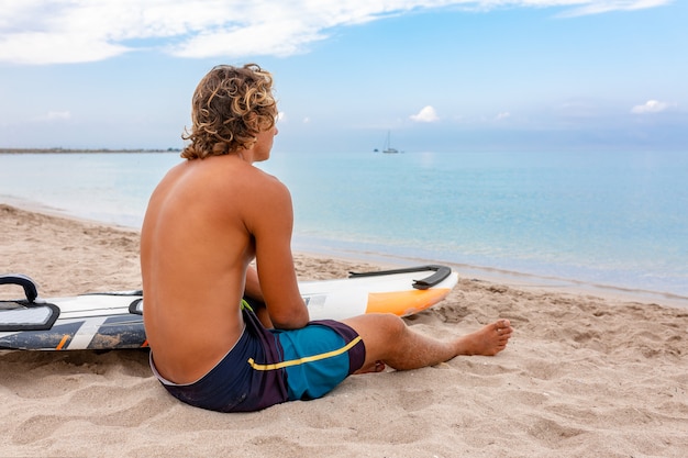 ハンサムな男が波に海の海の海岸でスポットをサーフィンするのを待つ白い空白のサーフィンボードとビーチに座っています。