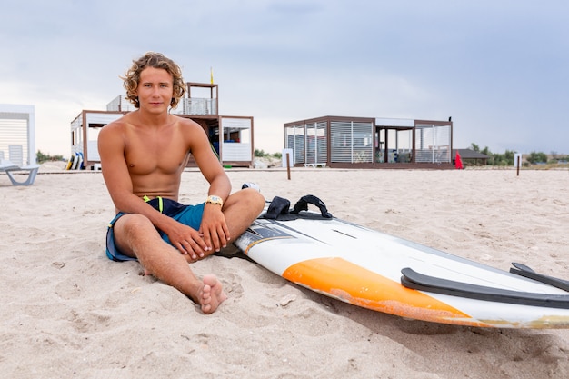 ハンサムな男が波に海の海の海岸でスポットをサーフィンするのを待つ白い空白のサーフィンボードとビーチに座っています。