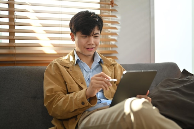 편안한 소파에 앉아 있는 잘생긴 남자는 태블릿에서 인터넷 검색을 즐깁니다. 사람과 기술 개념