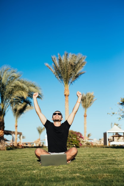 Handsome man sitting on the grass and use laptop with victory raised hands.