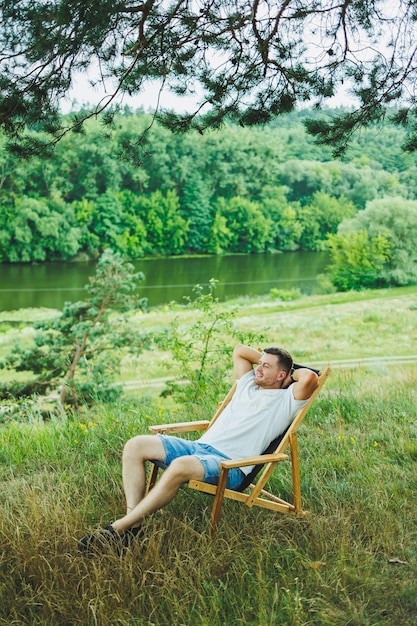 写真 木の椅子に座って自然の中で一人で座っているハンサムな男性 若いハンサムな男性が木の影のベンチに座って晴れた日に周囲の自然を楽しんでいます