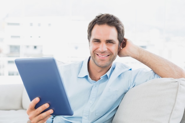 Handsome man sitting on the sofa using his tablet pc smiling at camera