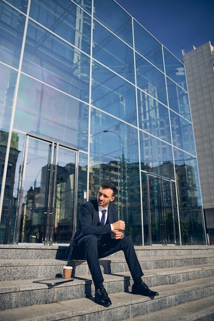 Handsome man sitting on stairs with coffee and relaxing near business center