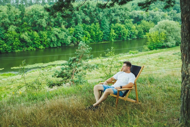 Handsome man sitting on a wooden chair in nature alone Young handsome guy sitting on a bench in the shade of trees and enjoying the surrounding nature on a sunny day