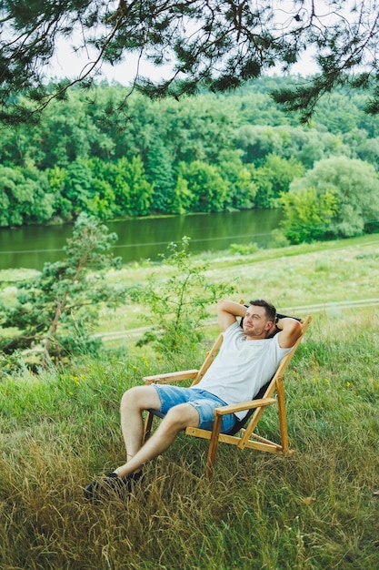 Handsome man sitting on a wooden chair in nature alone Young handsome guy sitting on a bench in the shade of trees and enjoying the surrounding nature on a sunny day