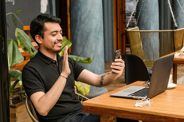 Handsome man smiling and making a cell phone video call and a computer and headphones on the table.