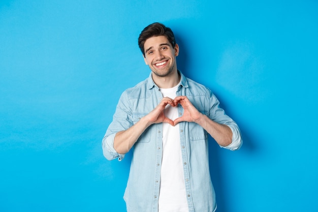 Handsome man smiling, showing heart gesture and looking at camera, saying I love you, standing against blue background