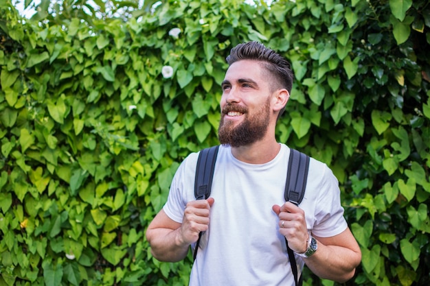 Handsome man smiling, with his adventure backpack on a background of green and natural leaves.