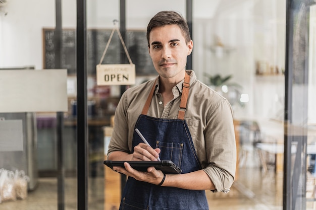 タブレットを持ってメモを取りながら立っているハンサムな男性は、店員であり、エプロンを着て、カフェでサービスを利用する顧客にサービスを提供しています。カフェサービスのコンセプト。