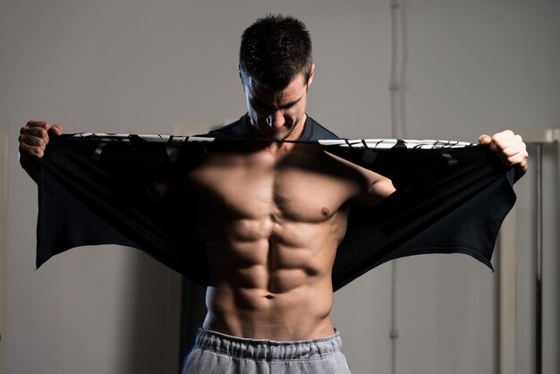 Красивый мужчина, стоящий крепко в спортзале и срывающий футболку с мускулистой спортивной бодибилдерской фитнес-модели, позирующей после упражнений
