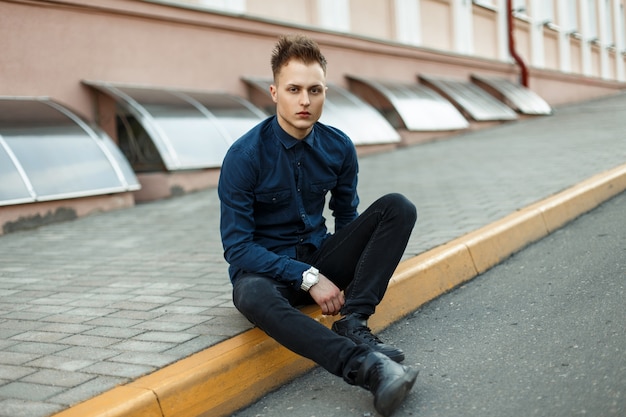 Handsome man in stylish clothes in black shoes sits on the asphalt