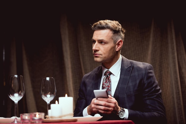 Handsome man in suit sitting at table and using smartphone in restaurant