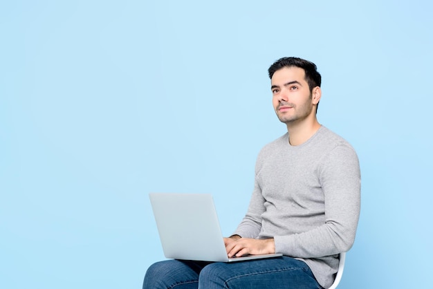 Handsome man thinking and looking up while using laptop computer on isolated light blue background