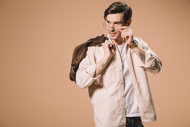 Handsome man touching glasses while standing with bag isolated on beige