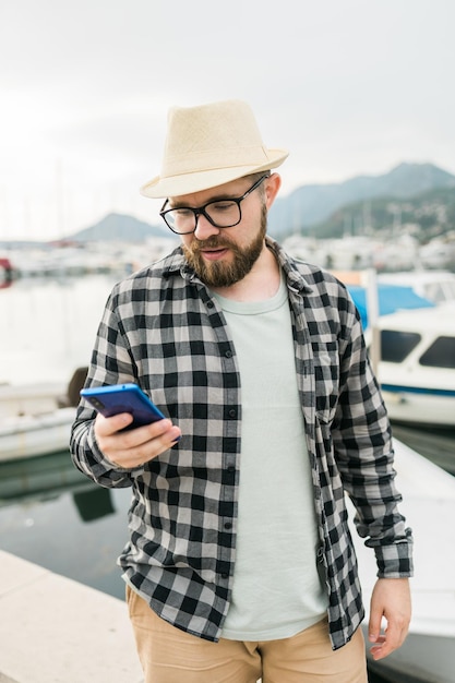 Handsome man tourist check in social networks during summer\
walk among sea marina with boats and yacht background traveller and\
social media app concept