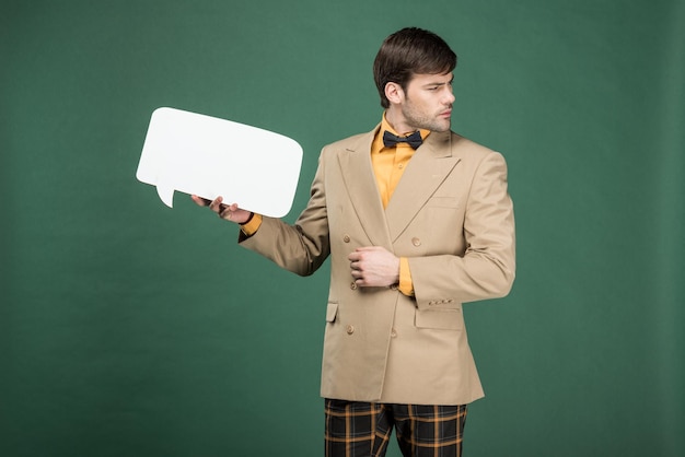 Handsome man in vintage clothes holding speech bubble and looking away isolated on green