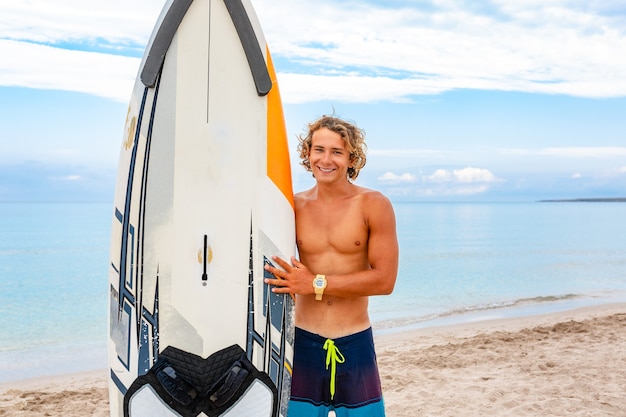 L'uomo bello cammina con la tavola da surf in bianco bianca aspetta che l'onda faccia surf sul punto del mare