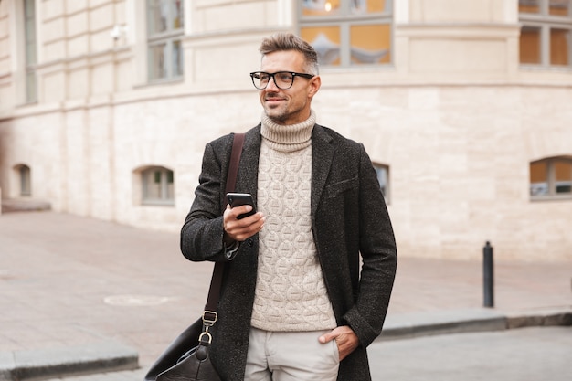 Handsome man wearing a coat walking outdoors, holding mobile phone