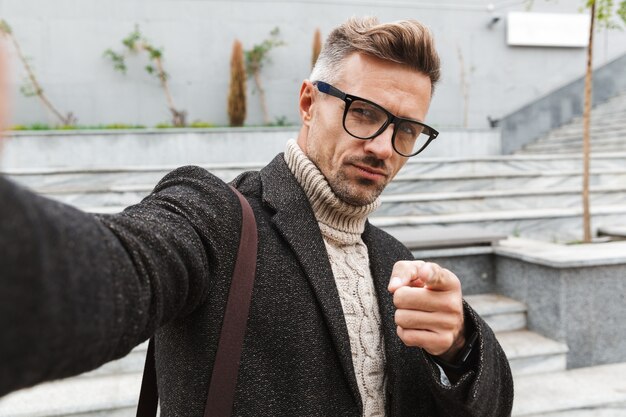 Handsome man wearing a coat walking outdoors, taking a selfie