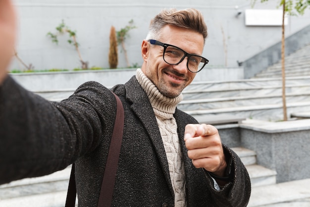 Handsome man wearing a coat walking outdoors, taking a selfie