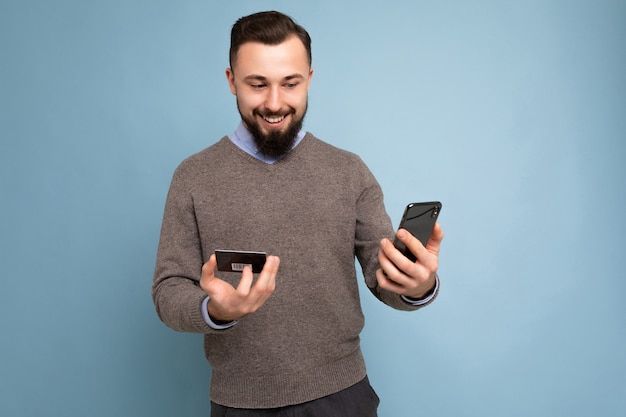 Handsome man wearing everyday clothes isolated on background wall holding and using phone and credit card making payment looking at smartphone screen.