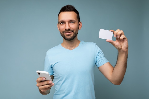 Handsome man wearing everyday clothes isolated on wall holding and using phone and credit card making payment looking at camera.