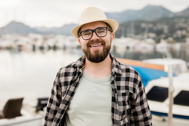 Handsome man wearing hat and glasses near marina with yachts Portrait of laughing man with sea port background