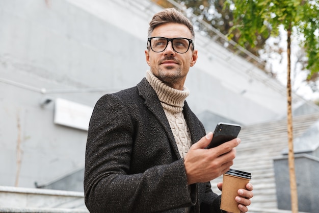 Handsome man wearing jacket holding mobile phone, drinking takeaway coffee while sitting outdoors