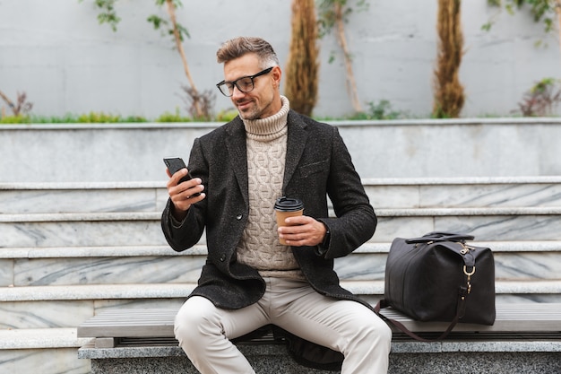 Handsome man wearing jacket holding mobile phone, drinking takeaway coffee while sitting outdoors