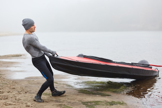 Handsome man wearing sporty wear and cap pulling kayak to bank from water