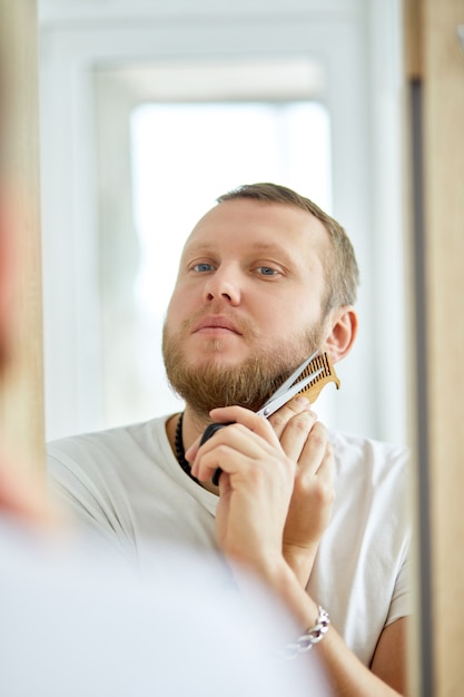 Handsome man in white t-shirt cutting beard himself with scissors at home