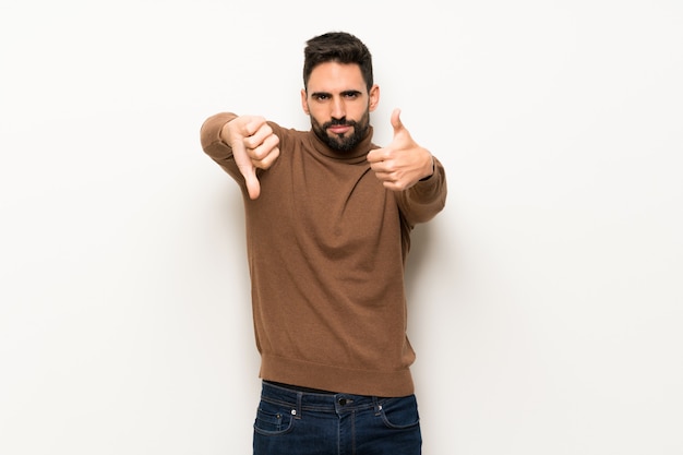 Handsome man over white wall making good-bad sign. Undecided between yes or not