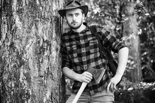 Photo handsome man with axe handsome man lumberjack with a large ax examines the tree before felling strong man lumberjack with an ax in a plaid shirt