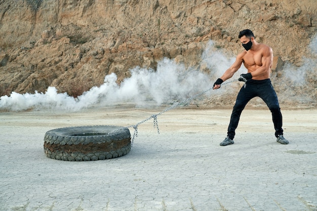 Handsome man with bare muscular torso, wearing sport pants and medical mask pulling heavy tyre that fixed on long metal chain. Concept of strength and endurance.